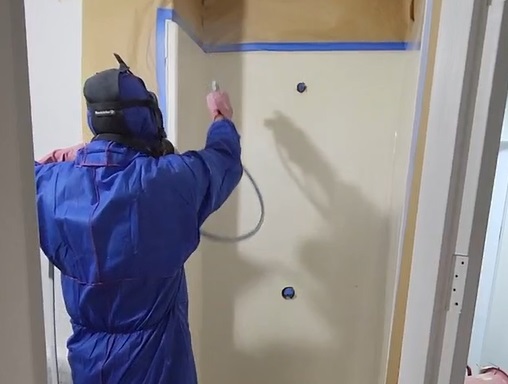 A person in blue coveralls painting the wall of a room.